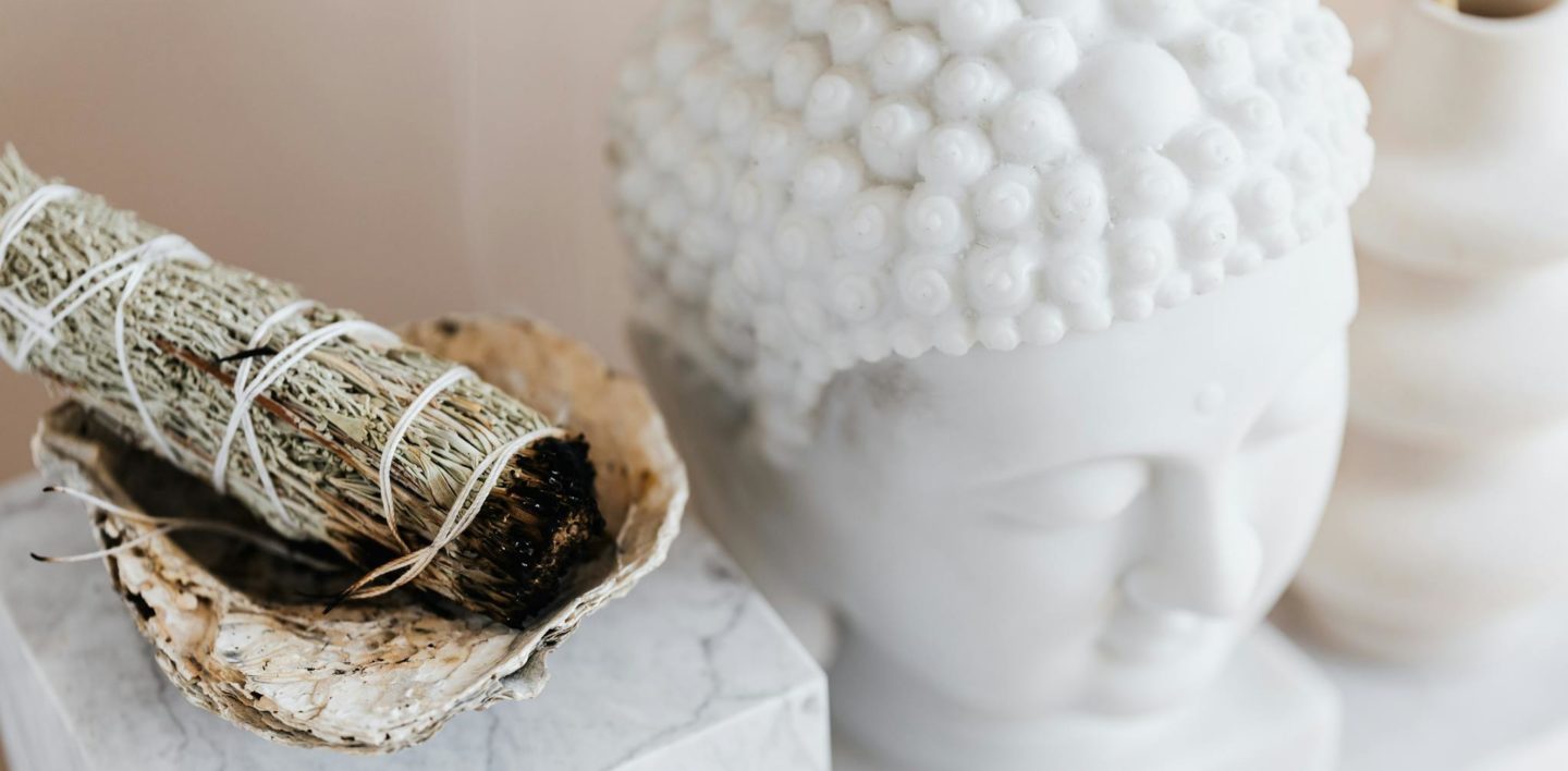 From above of sage candle in bowl placed on white marble shelf near plaster Buddha bust and creative shape vase for home decoration
